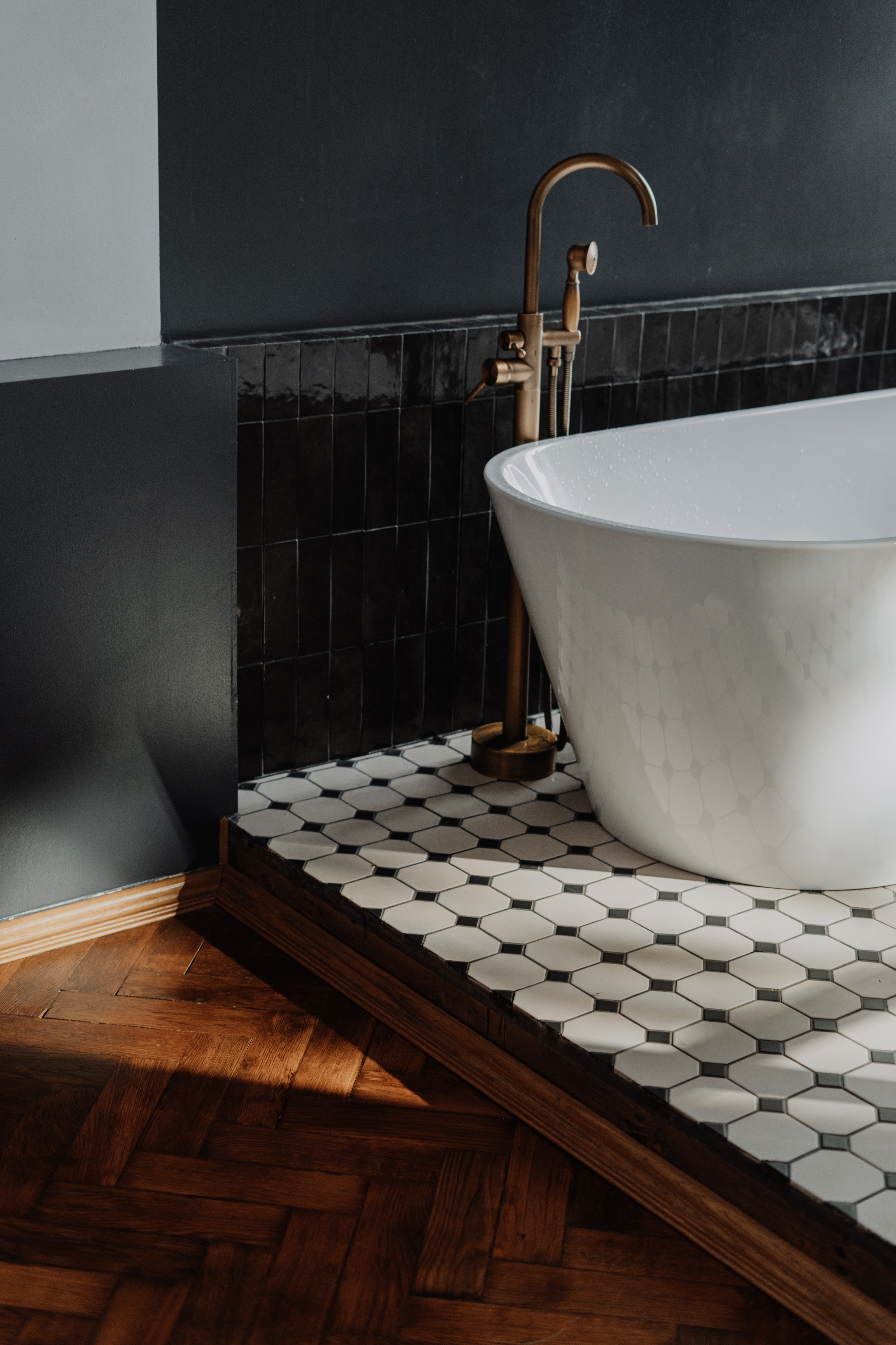 Vertical stacked gloss black metro tiles sit behind a modern freestanding bathtub. Underneath it are stylish back and white mosaic floor tiles in a geometric shape.